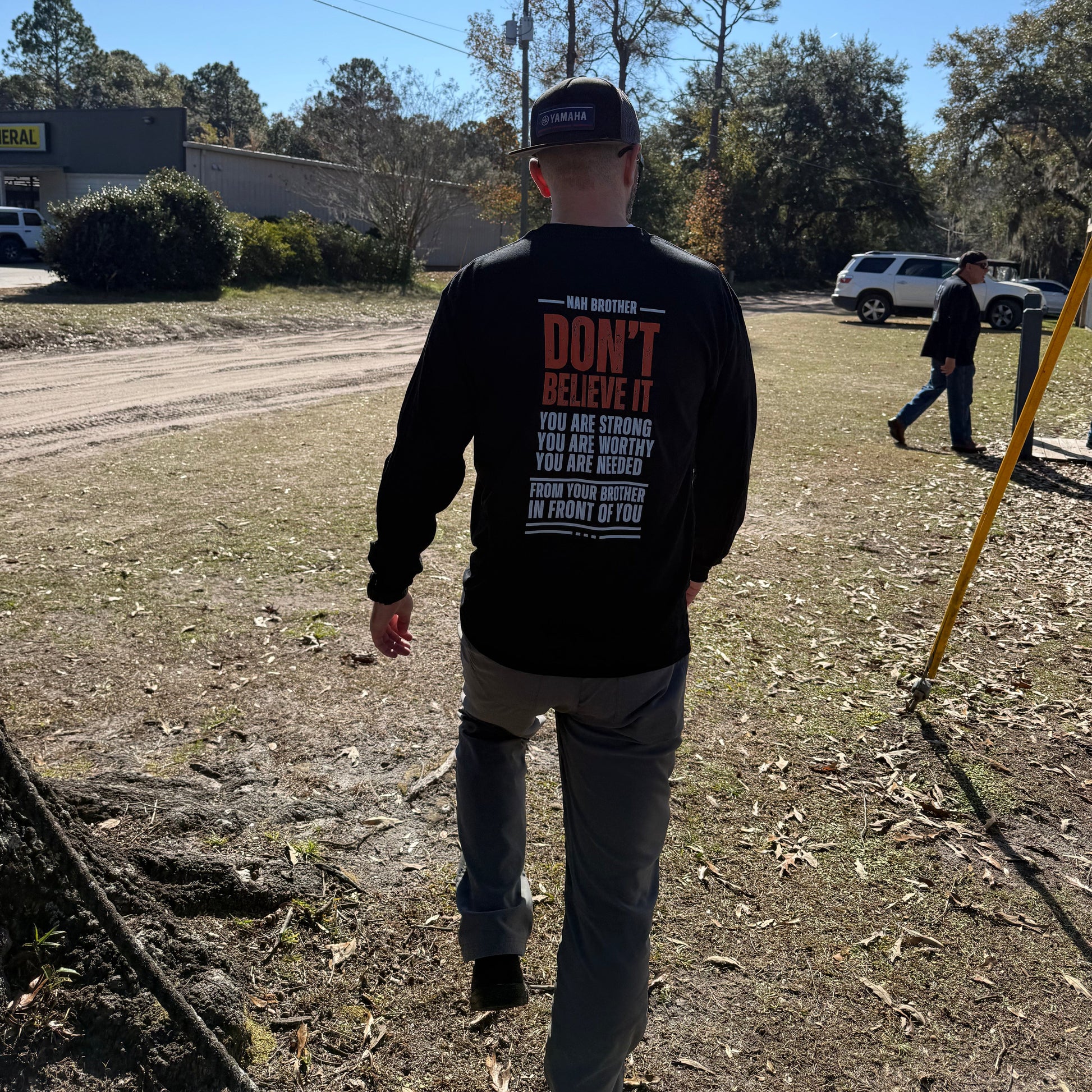 Person wearing a black shirt with text, standing on a dirt road with trees and a blue sky in the background.