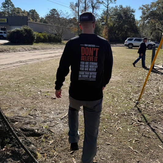 Person wearing a black shirt with text, standing on a dirt road with trees and a blue sky in the background.