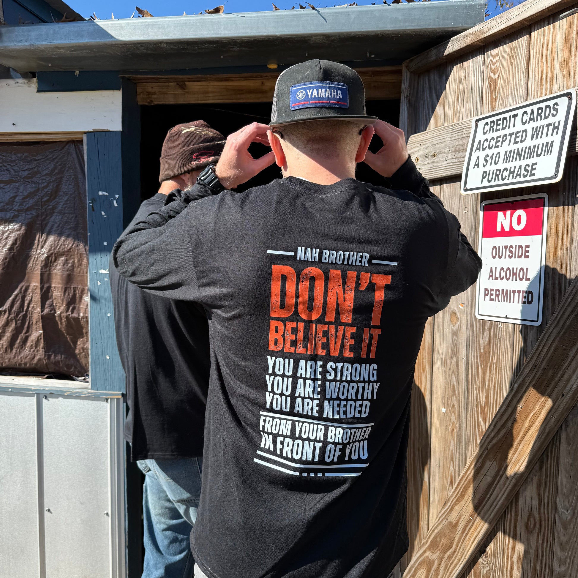 Person wearing a black shirt with motivational text, standing outdoors near wooden structures.