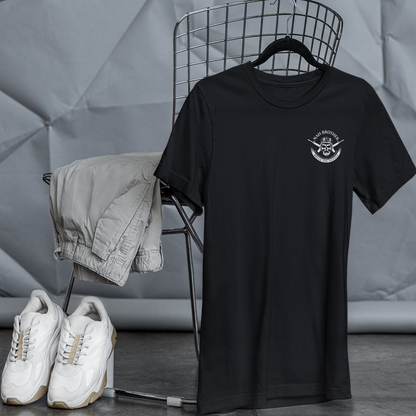 Black t-shirt with a logo on a hanger, white sneakers on the floor, and folded beige jacket on a chair against a gray textured wall.